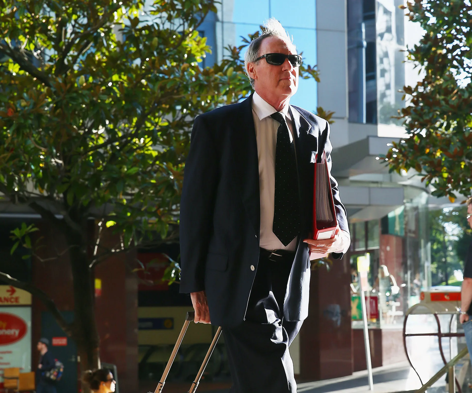 Man in suit and sunglasses walks outside with documents and briefcase in an urban setting.