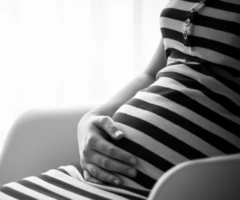 Pregnant woman in striped dress cradling belly, seated, in grayscale.