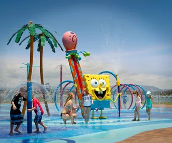 Children playing in a water park featuring a large SpongeBob statue with water sprays and palm tree designs.