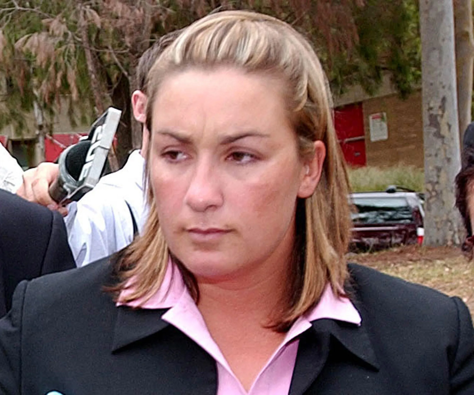 A woman outdoors wearing a black blazer over a pink shirt, looks pensive with people around her.