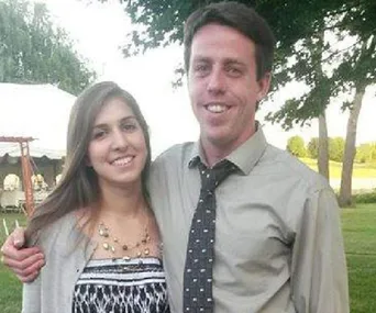 A smiling couple posing outdoors, with a tent and trees in the background.