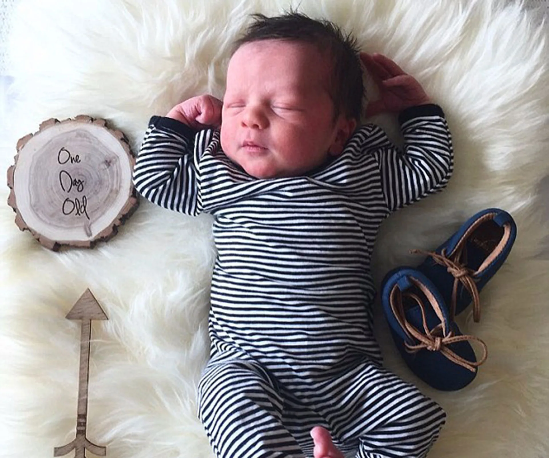 Newborn baby in striped outfit lying on soft fur, with sign reading "One Day Old" and blue moccasins beside him.