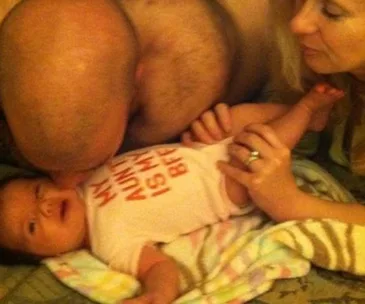 A baby in a onesie lies on a bed with a person kissing and another holding the baby's leg; an optical illusion creates confusion.