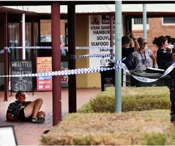 A man sits on the ground outside a shop, surrounded by police tape. Officers and onlookers are near a storefront.