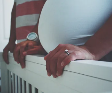 A pregnant belly with hands resting on a crib rail, one wearing a ring.