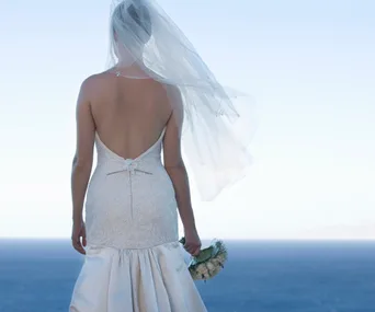 Back view of a bride in a lace gown and veil, holding flowers by the ocean.