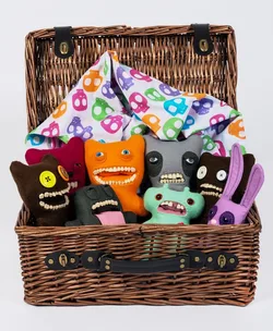 A wicker basket holds colorful plush dolls with quirky expressions and teeth, set against a skull-patterned cloth.