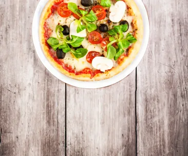 Pizza with mushrooms, tomatoes, olives, greens on white plate on wooden table background.