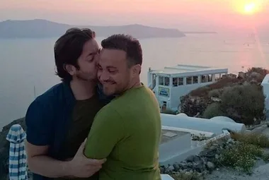 Two men embrace and share a moment during sunset on a coastal hillside, with a view of the sea and distant islands.