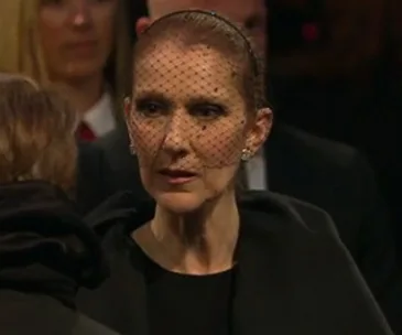 A woman in a black outfit and veil attends a solemn event, appearing emotional and focused.