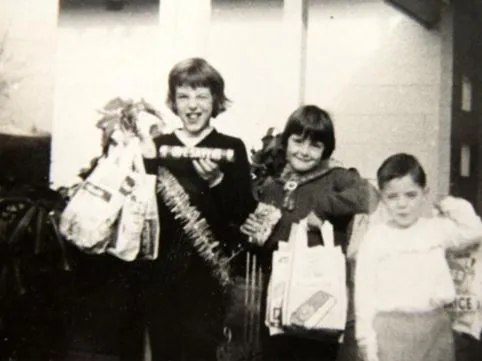 Three children standing together, smiling, holding shopping bags and a wrapped box in a black-and-white photo.