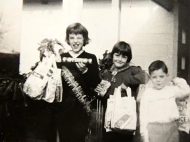 Three children standing together, smiling, holding shopping bags and a wrapped box in a black-and-white photo.