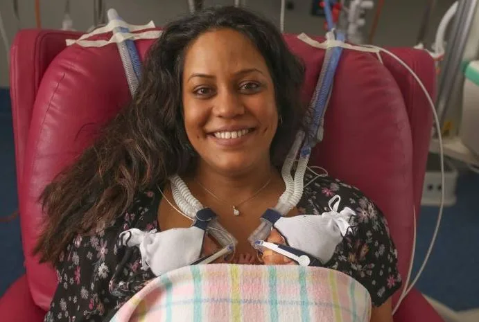 Smiling woman sits in hospital chair for skin-to-skin contact with premature twins, tubes attached for support.