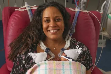 Smiling woman sits in hospital chair for skin-to-skin contact with premature twins, tubes attached for support.