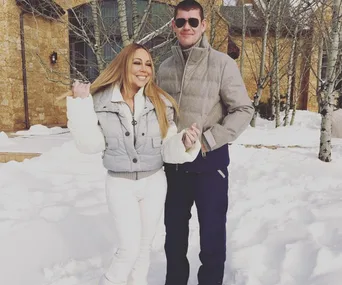 Couple standing in a snowy outdoor setting, with trees and a building in the background, both wearing winter clothes.