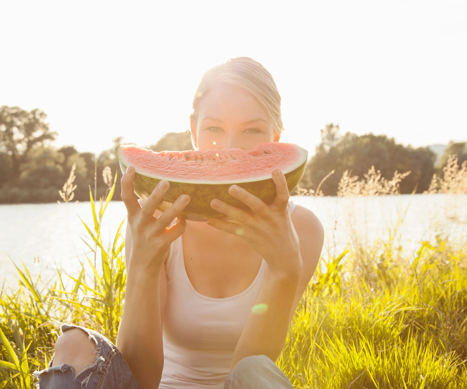 Woman in sunlight sitting by a lake, holding a large slice of watermelon near her face.