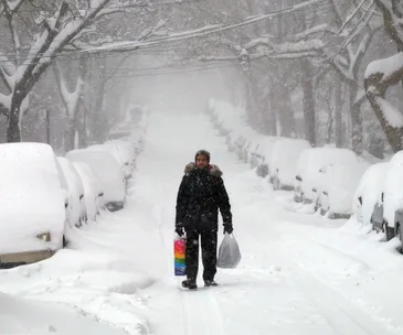 A person walks down a snow-covered street carrying shopping bags during heavy snowfall.