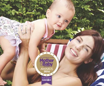 A mother smiling while holding her baby outdoors, featured in Australian Mother & Baby Awards 2016 with green foliage in background.