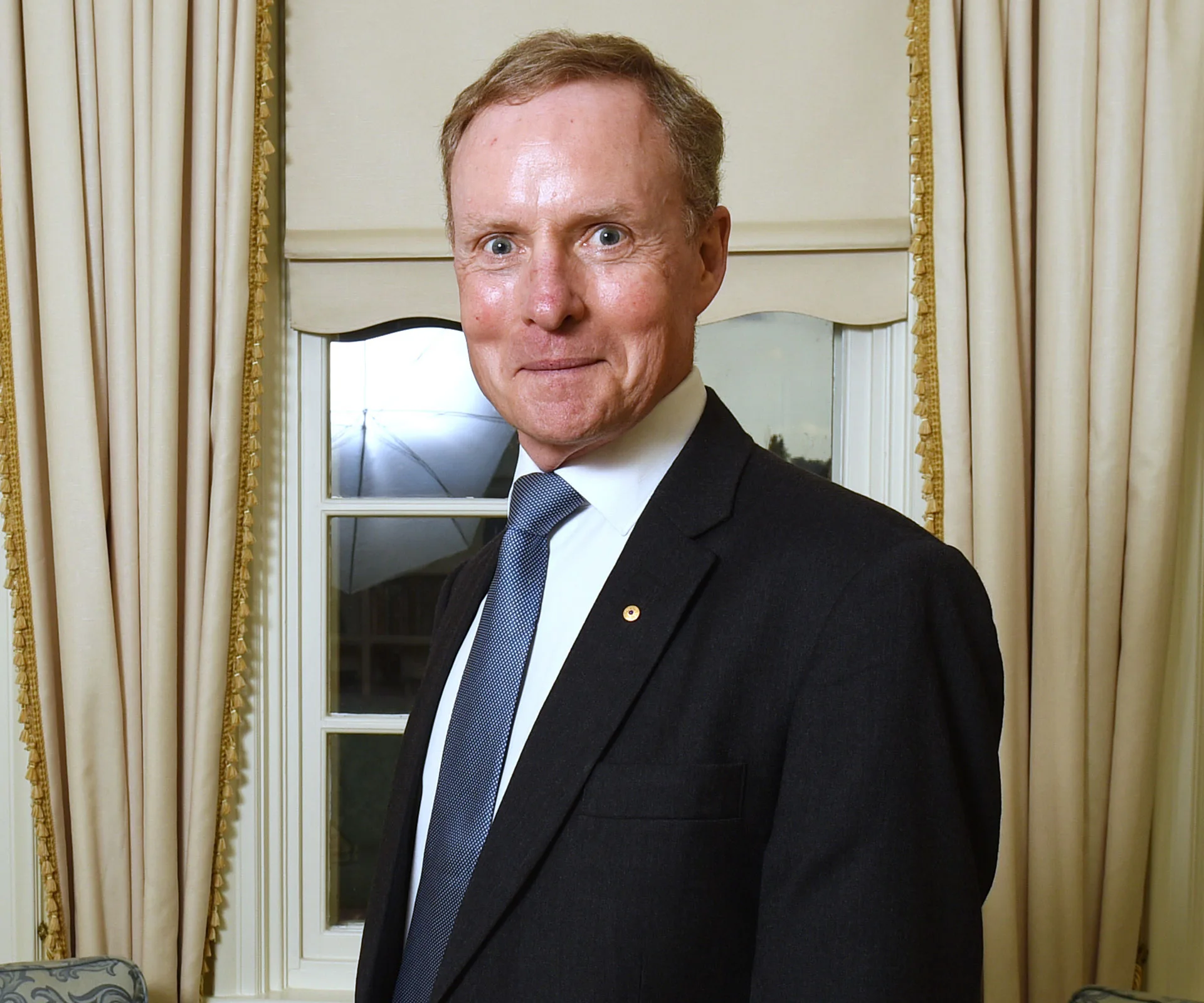 Man in a suit standing in a room, smiling, with curtains and a window in the background.