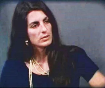 A woman with long dark hair wearing earrings and a necklace, seated indoors against a plain background.