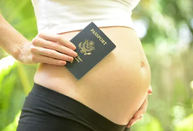 Pregnant woman holding a U.S. passport against her belly, outdoors.