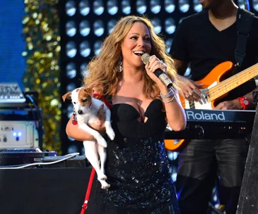 Mariah Carey holds a small dog on stage, singing with musicians in the background.