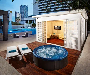 Luxury poolside cabana with loungers, a hot tub, and modern city skyline in the background at dusk.