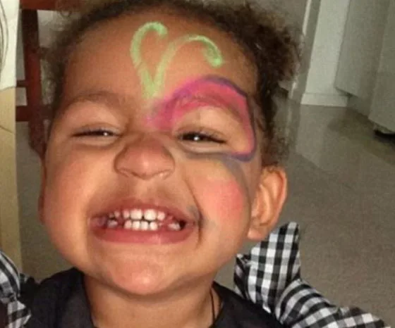 Toddler smiling with colorful face paint, eyes closed, wearing a checkered top.