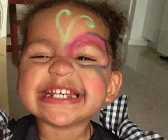 Toddler smiling with colorful face paint, eyes closed, wearing a checkered top.