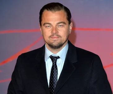 A man in a suit and tie stands against a red and purple backdrop, looking directly at the camera.