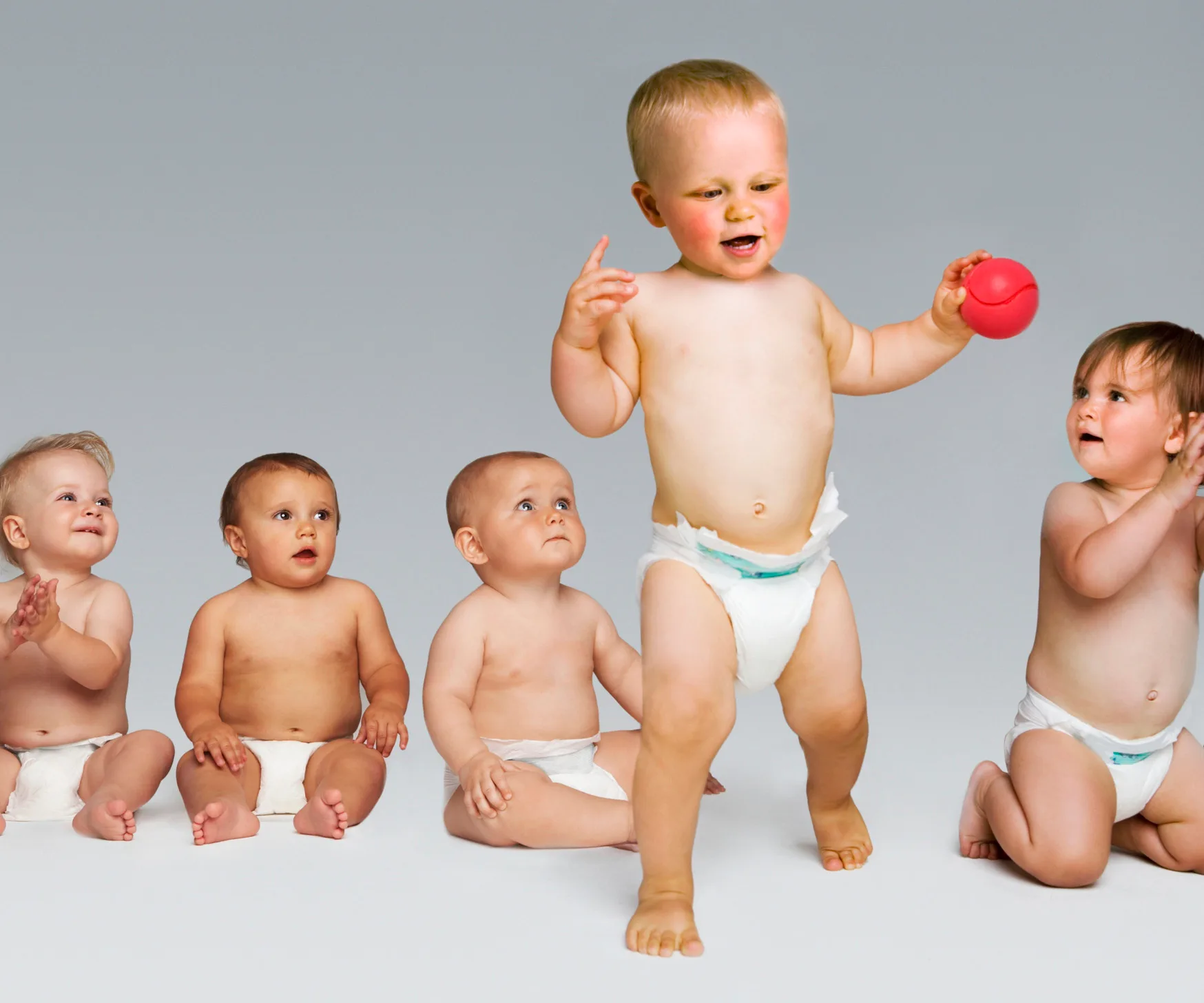 Five babies in diapers, sitting and one standing, holding a red ball against a neutral background.