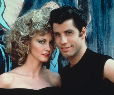 A scene from "Grease" with Sandy and Danny, dressed in 1950s-style outfits, standing close together.