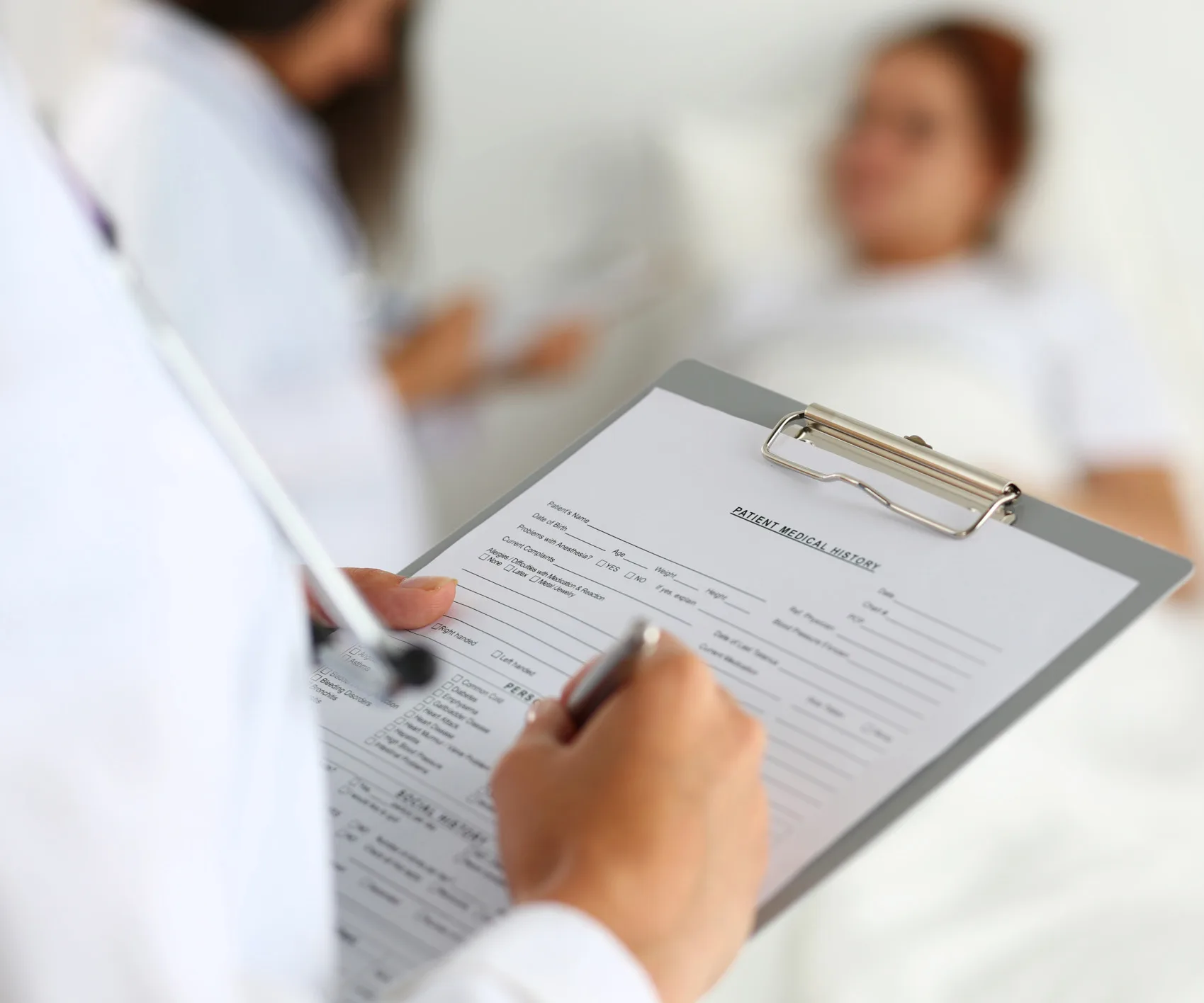 Doctor holding a clipboard near a patient lying in a hospital bed, with medical forms visible.