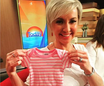 Woman smiling, holding a pink striped baby outfit on a TV set with "Today" on the screen in the background.