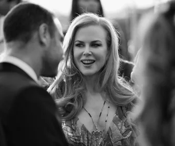 A woman with long hair and a necklace smiles while talking to a man in a formal setting, captured in black and white.