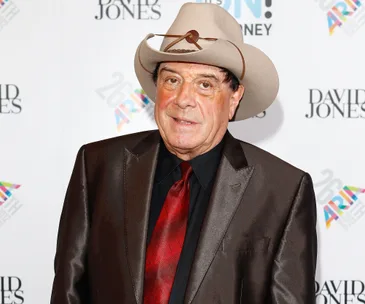 A man in a cowboy hat and suit with a red tie stands in front of a backdrop with colorful logos.