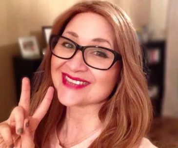 A woman with long hair and glasses smiles, making a peace sign with her fingers in a warmly lit room.