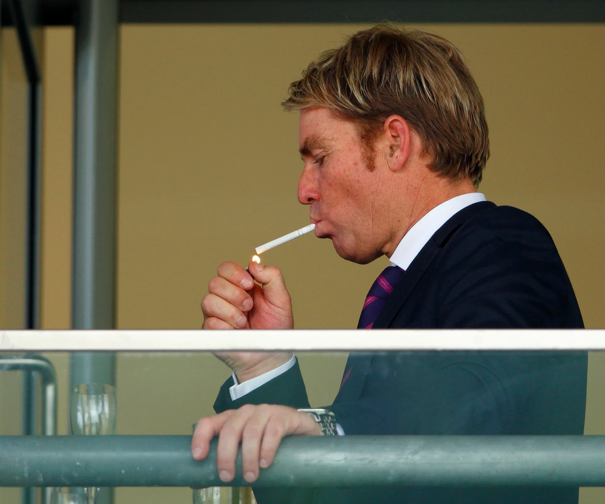 Man in a suit lighting a cigarette on a balcony.