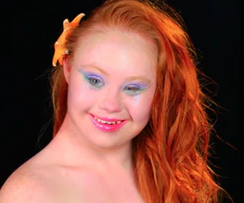 Red-haired woman with an orange starfish hair clip, vibrant makeup, smiling against a black background.