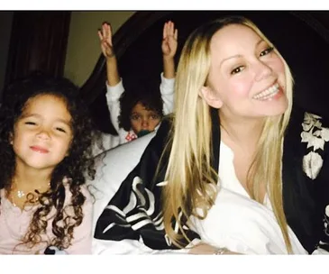 Mariah Carey smiling with her kids on a bed, one child making a playful gesture in the background.