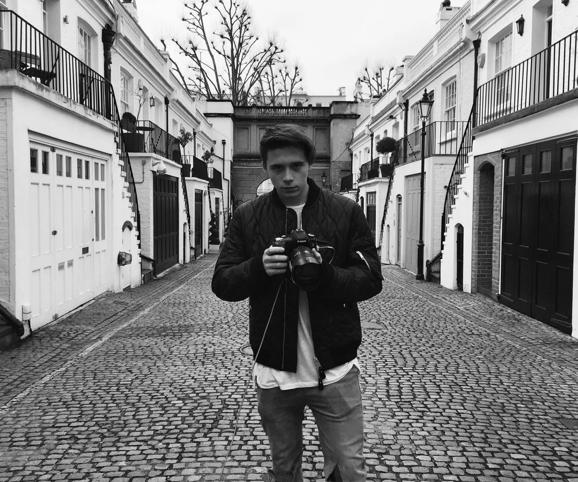 Young man holding a camera stands on a cobblestone street between white townhouses on an overcast day.