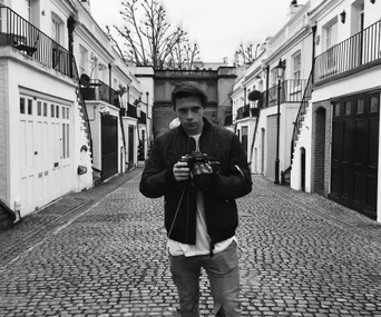 Young man holding a camera stands on a cobblestone street between white townhouses on an overcast day.