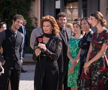 Sophia Loren, in black lace, holds a rose surrounded by people, in an outdoor setting.
