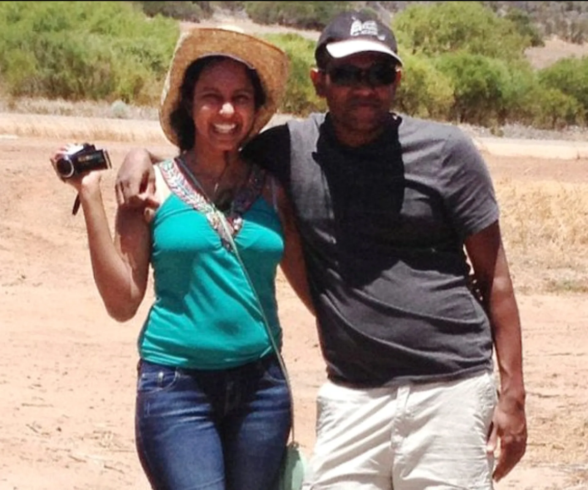 A couple stands together outdoors, the woman in a hat holding a camcorder, both smiling in sunny weather.