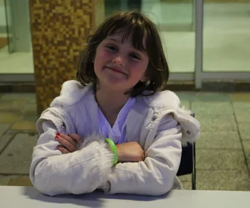 Young girl smiling, wearing a white coat and green wristband, seated with arms crossed at an outdoor table.