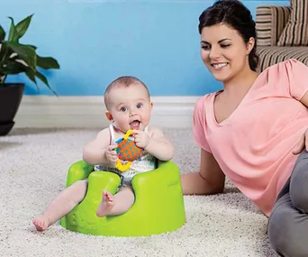 Baby in a green seat playing with a toy, smiling woman beside on carpet.