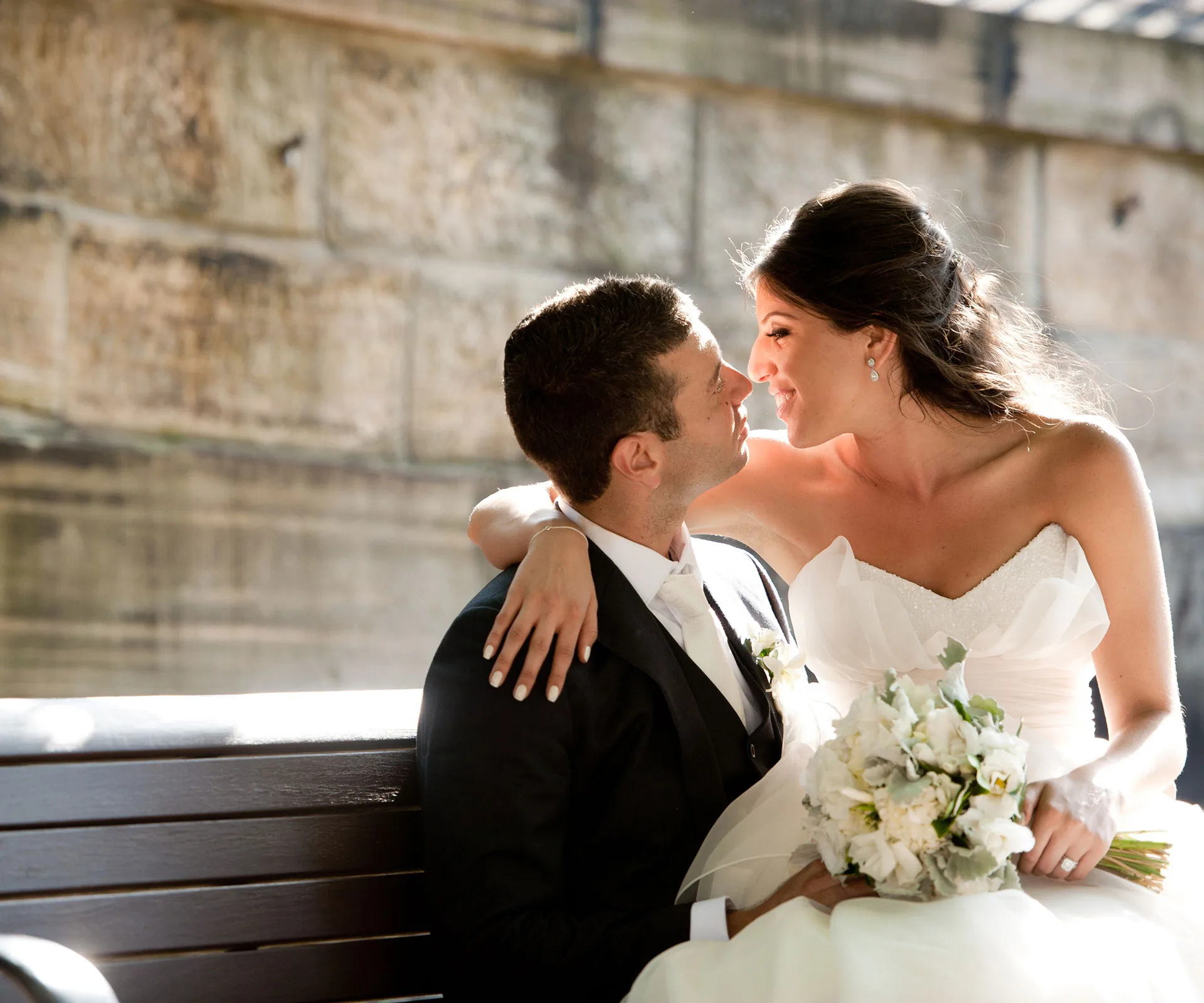 A bride and groom sitting on a bench, smiling and embracing, with the bride holding a bouquet of white flowers.