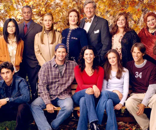 Cast of "Gilmore Girls" gathered in front of autumn leaves, featuring multiple characters from the show smiling together.