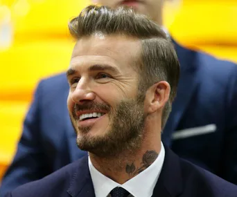 A smiling man with a beard and styled hair, wearing a suit and sitting in a brightly lit setting.