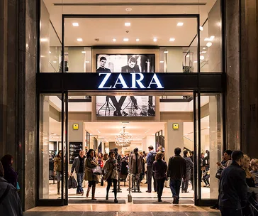 Shoppers entering a busy Zara store with bright signage and fashion poster above in a city setting.